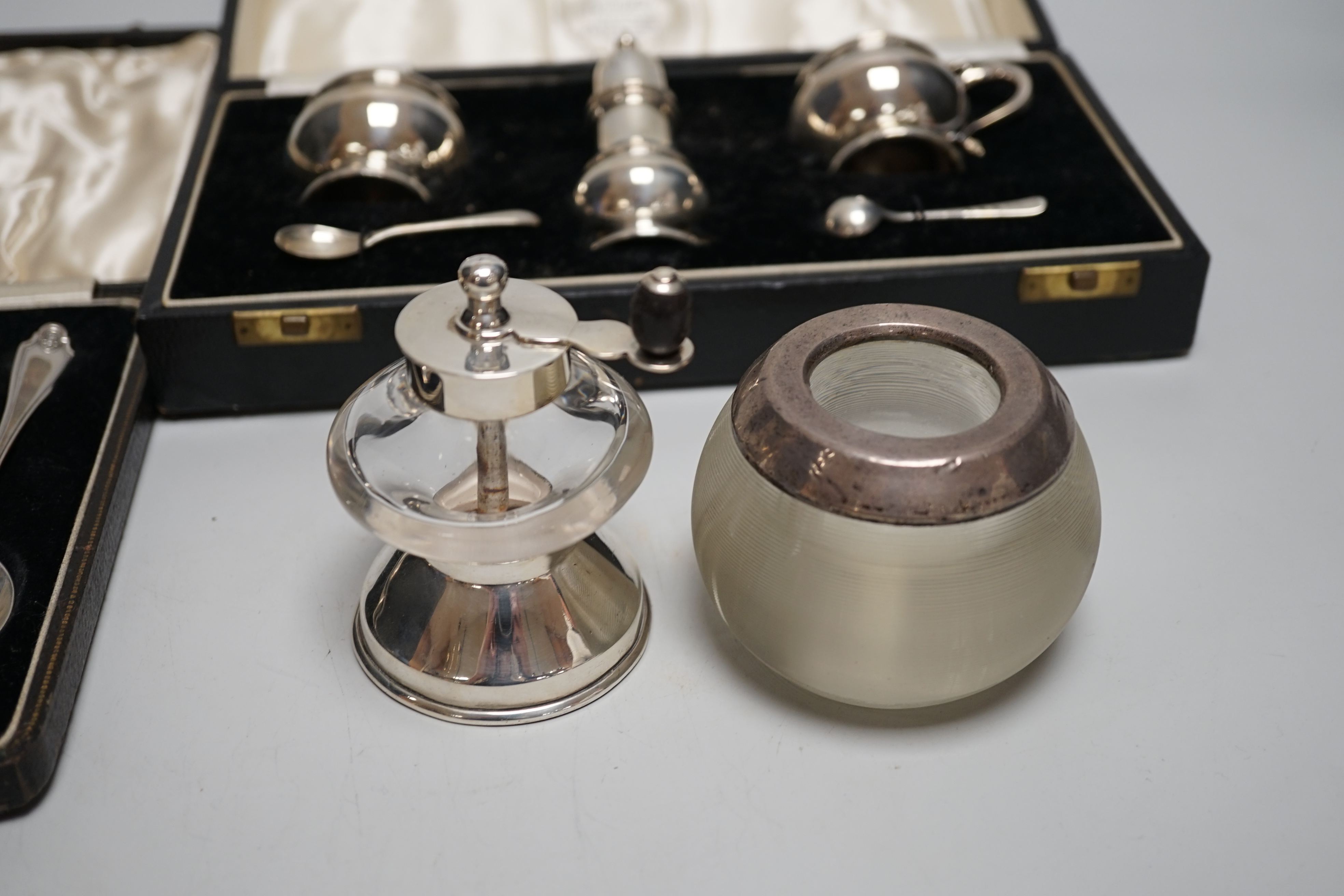 A cased George V silver three piece condiment set with two spoons, London, 1935, a cased set of six silver grapefruit spoons, a silver mounted glass pepper grinder and a white metal mounted glass match strike.
